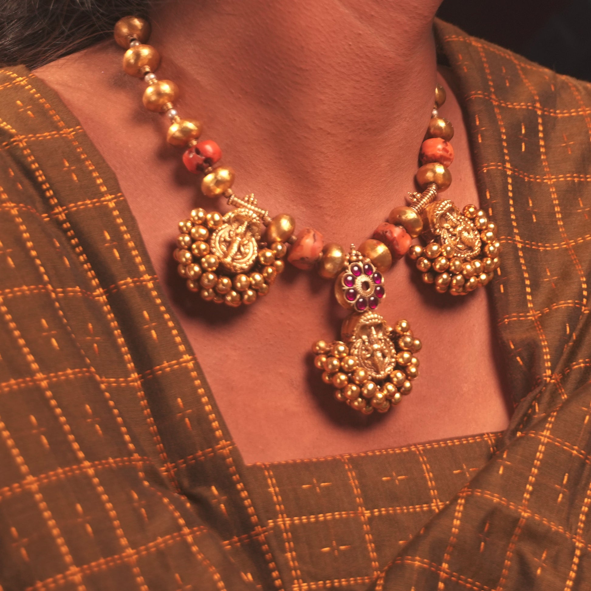 A close-up of a person wearing an ornate gold-toned traditional necklace with decorative motifs and coral-colored beads, paired with a brown patterned sari.