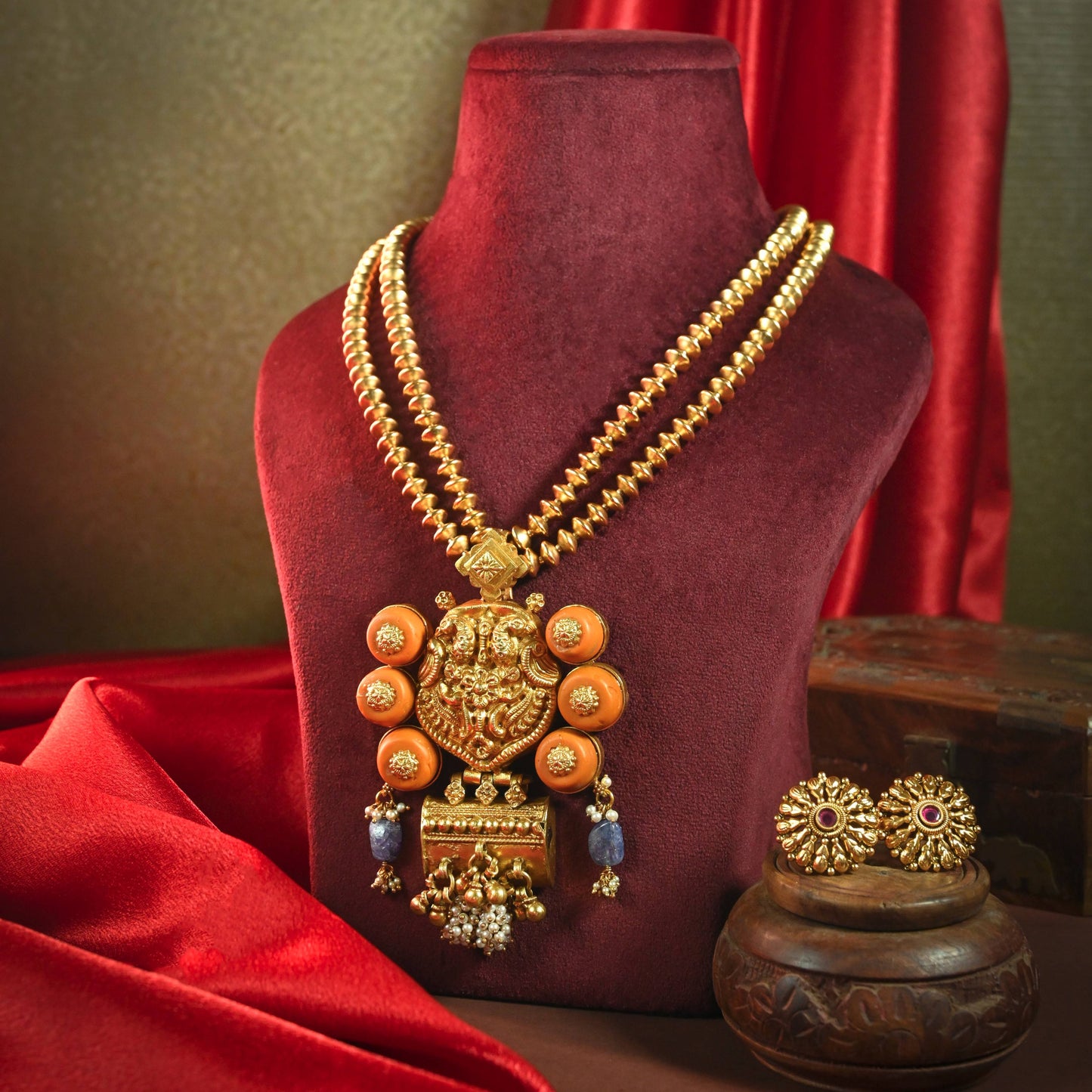 an ornate gold-toned traditional necklace displayed on a dark red velvet mannequin bust, with matching earrings and a decorative pot beside it against a draped red fabric and olive background.