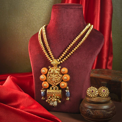 An ornate gold-toned traditional necklace displayed on a dark red velvet mannequin bust, with matching earrings and a decorative pot beside it against a draped red fabric and olive background.